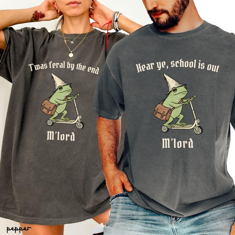 May include: Two dark grey t-shirts with a frog graphic riding a scooter. One shirt reads "Twas feral by the end, M'lord" and the other says "Hear ye, school is out, M'lord". The frog wears a hat and carries a satchel.