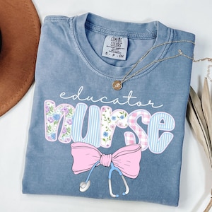 May include: A light blue t-shirt with the text "educator nurse" in a patchwork style with floral and plaid patterns. A pink bow with a stethoscope is below the text.