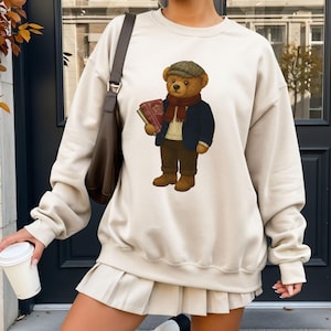 May include: Cream-colored sweatshirt featuring a cartoon bear wearing a tweed cap, scarf, and jacket, holding books. The bear is depicted in a classic, collegiate style. The sweatshirt is paired with a pleated skirt and a brown shoulder bag.