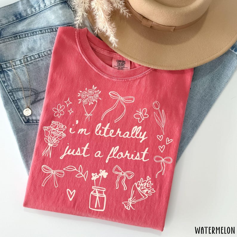 May include: A watermelon-colored t-shirt with the text "I'm literally just a florist" in white script. The shirt is decorated with floral illustrations, bows, and hearts. A tan hat and blue jeans are also in the image.