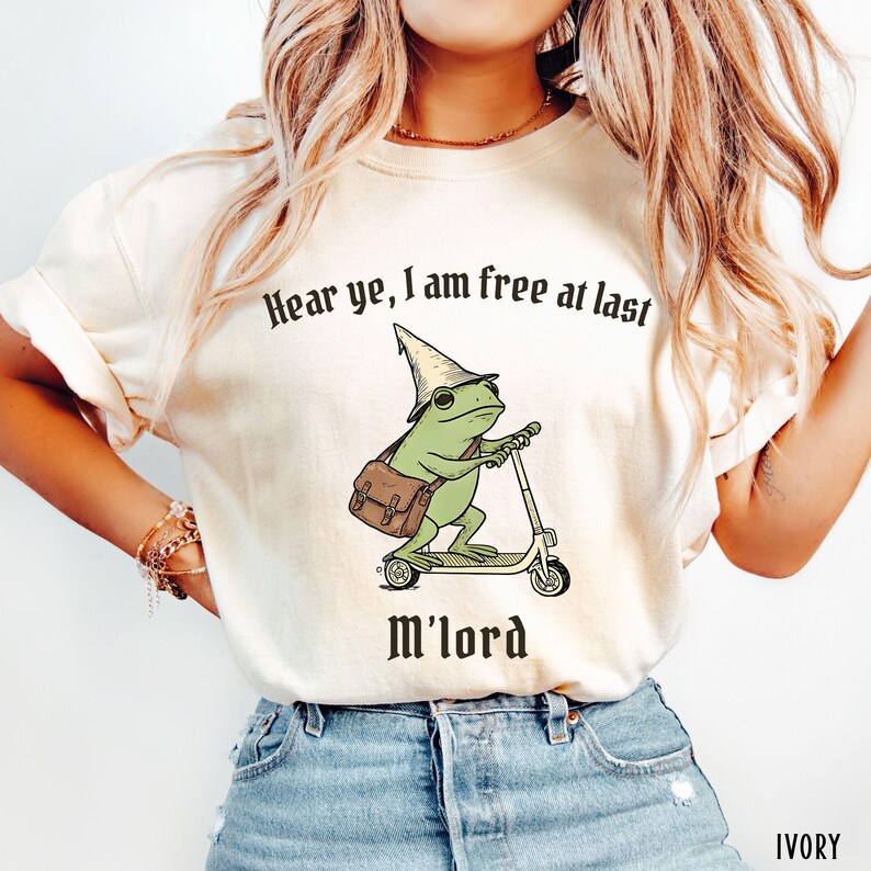 May include: Ivory-colored t-shirt featuring a green frog wearing a wizard hat and carrying a satchel, riding a scooter. The text "Hear ye, I am free at last M'lord" is printed above and below the frog illustration.