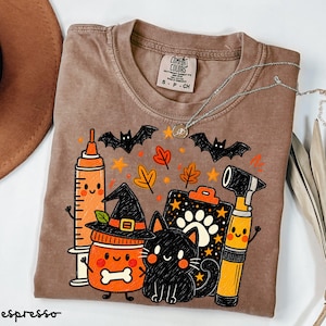 May include: A brown short-sleeved t-shirt featuring a graphic print of a syringe, a jar wearing a witch hat, a black cat, and an otoscope. The design includes orange autumn leaves, stars, and bats. The inner neck label reads Comfort Colors.