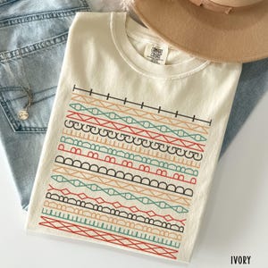 May include: Ivory-colored t-shirt with a colorful geometric design. The design features horizontal rows of various patterns in red, green, orange, and black. The shirt is made of a soft material and is folded on top of blue jeans.