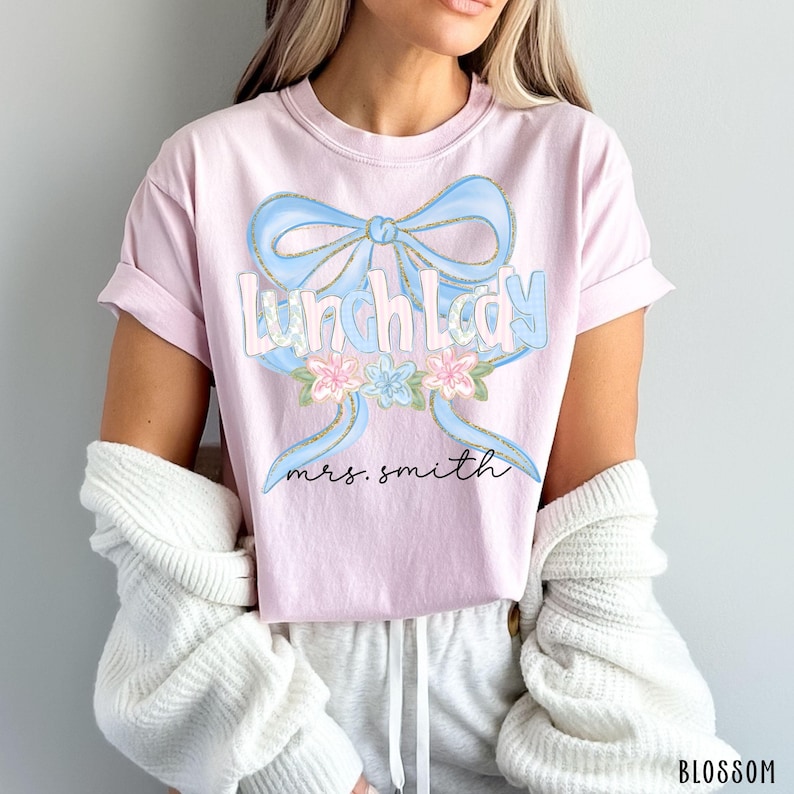 Puede incluir: Camiseta rosa claro con la frase "Lunch Lady" en azul claro, contorneada en dorado, y un dise&ntilde;o de lazo azul. La camiseta tambi&eacute;n presenta el nombre "mrs. smith" y un dise&ntilde;o floral.