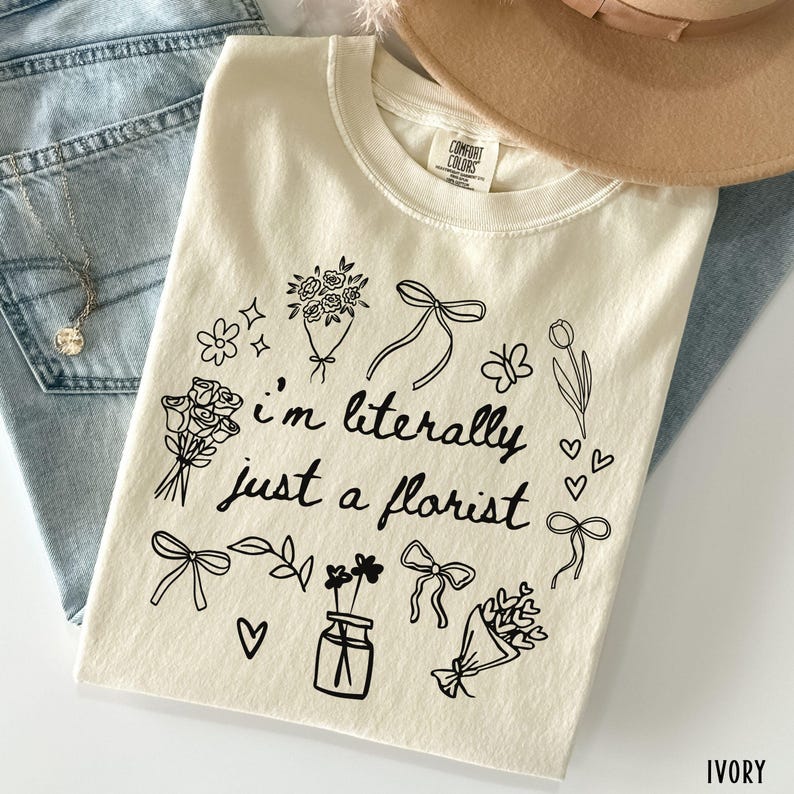 May include: Ivory-colored t-shirt with the text "I'm literally just a florist" surrounded by floral and bow illustrations. The shirt is made of a soft material and is folded on a light blue denim surface.