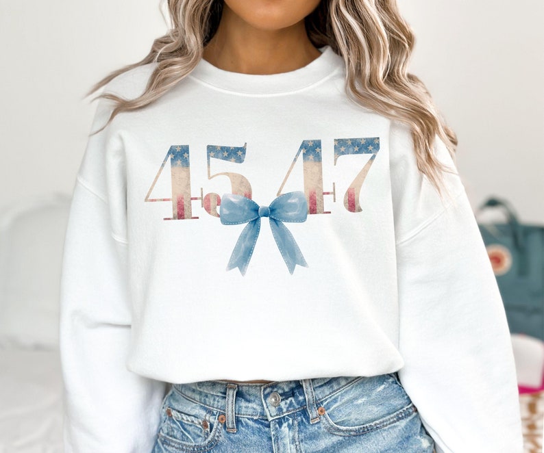 May include: White sweatshirt with the numbers 4547 printed in red, white, and blue, resembling the American flag. A blue bow is centered below the numbers.