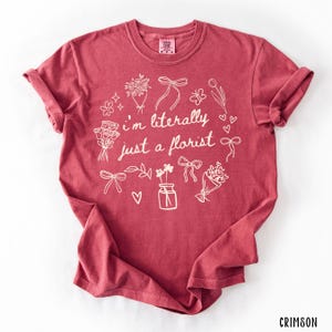 May include: Crimson-colored t-shirt with rolled-up sleeves. The shirt features a white floral design with the text "I'm literally just a florist". The design includes bouquets, bows, and a vase of flowers.