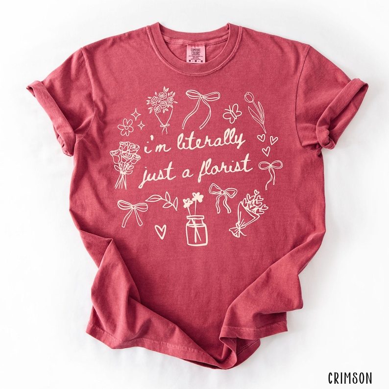 May include: Crimson-colored t-shirt with rolled-up sleeves. The shirt features a white floral design with the text "I'm literally just a florist". The design includes bouquets, bows, and a vase of flowers.