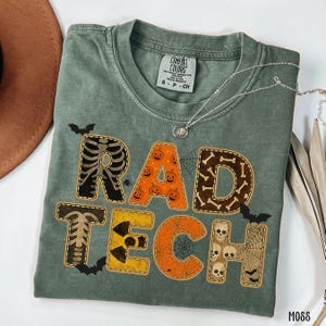May include: A moss green t-shirt featuring a Halloween design. The words "RAD TECH" are displayed with spooky graphics, including a skeleton, pumpkins, bones, skulls, and spiderwebs. The shirt is made of a soft material.