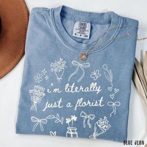 May include: A blue jean Comfort Colors t-shirt with the text "I'm literally just a florist" surrounded by floral illustrations. A gold necklace with a pendant rests on the shirt.