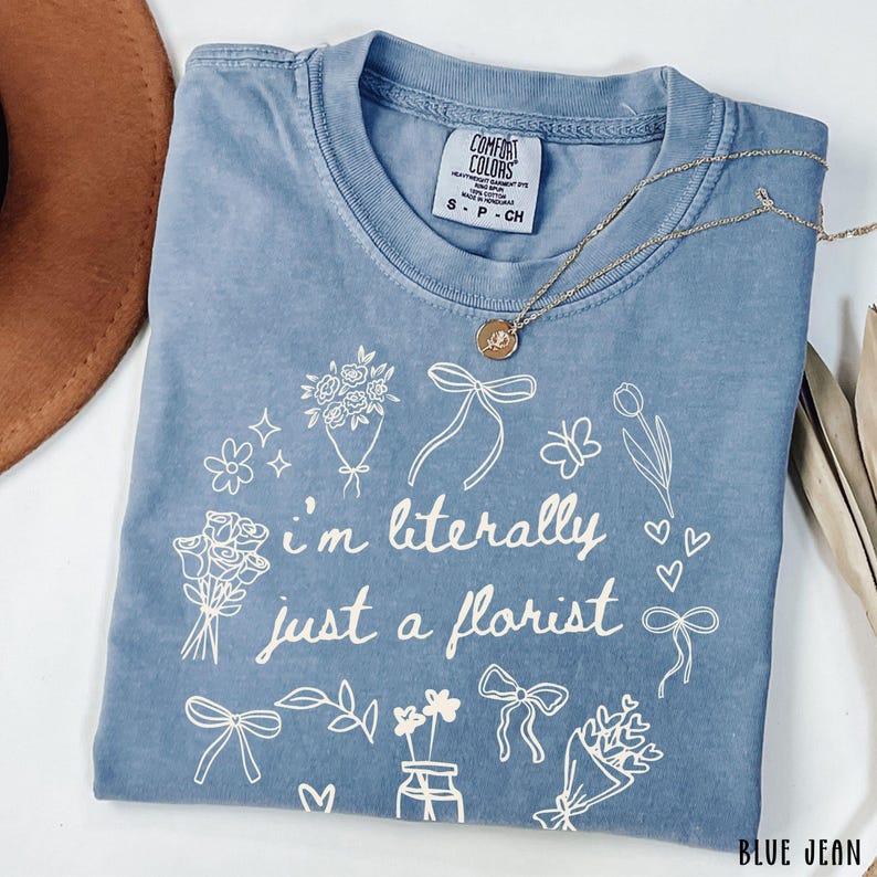 May include: A blue jean Comfort Colors t-shirt with the text "I'm literally just a florist" surrounded by floral illustrations. A gold necklace with a pendant rests on the shirt.