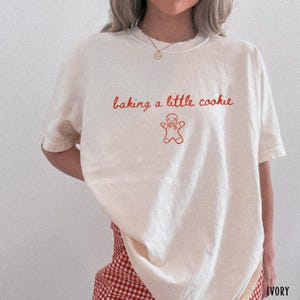 May include: An ivory short-sleeved t-shirt featuring the red text "baking a little cookie" and a red line-art illustration of a gingerbread person. The shirt has a crew neckline and is worn with red and white checkered bottoms.