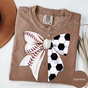 May include: A brown t-shirt with a white bow made from a baseball and a soccer ball. The shirt is a Comfort Colors brand and the color is Espresso.