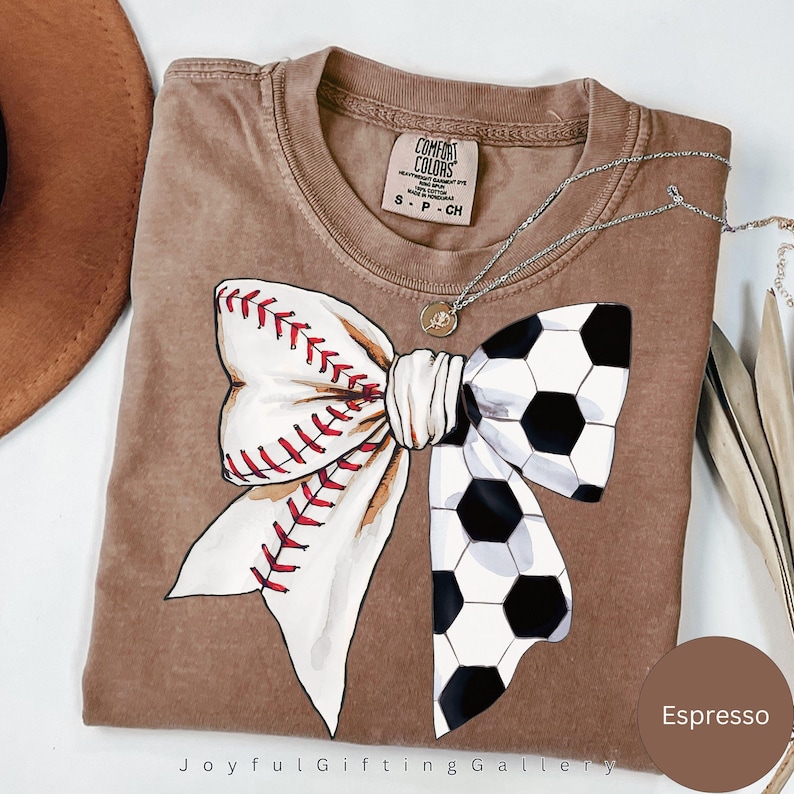 May include: A brown t-shirt with a white bow made from a baseball and a soccer ball. The shirt is a Comfort Colors brand and the color is Espresso.