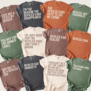 May include: A collection of eleven short-sleeved crew neck t-shirts in various colors including brown, charcoal, orange, and green. Each shirt features white or dark brown text with humorous phrases about deviled eggs, such as Deviled Egg Dealer.