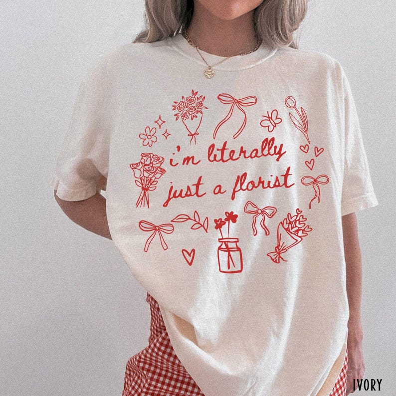 May include: Ivory-colored t-shirt with the text "I'm literally just a florist" in red. The design includes floral illustrations, bows, and hearts. The shirt is a casual, comfortable style.