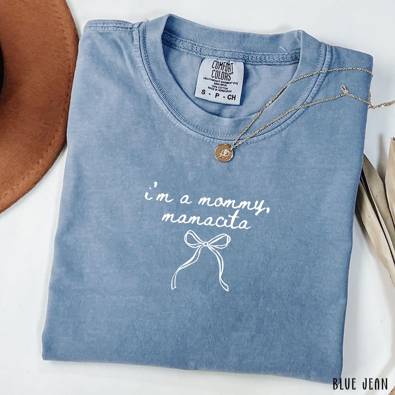 May include: A blue Comfort Colors t-shirt with the text "I'm a mommy, manacita" and a bow graphic. A gold necklace with a pendant is draped across the shirt. The shirt is folded and has a tag at the neckline.