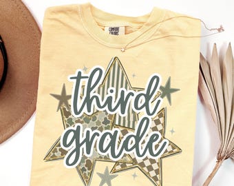 Può includere: Una t-shirt a maniche corte giallo pallido con un'etichetta Comfort Colors sul collo. La grafica presenta il testo "third grade" in corsivo grigio scuro, sovrapposto a tre grandi stelle con motivi a righe, mimetici e a scacchi.