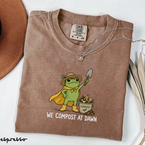 May include: A brown t-shirt featuring a graphic of a green frog wearing a yellow cape, a hat, and yellow boots. The frog holds a small shovel next to a bucket labelled COMPOST. The text WE COMPOST AT DAWN is printed below the graphic.