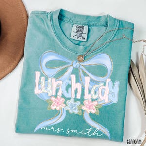Puede incluir: Camiseta verde mar con un lazo azul claro y las palabras "Lunch Lady" en una fuente pastel. La camiseta tambi&eacute;n presenta detalles florales y el nombre "mrs. smith" en cursiva. Un collar plateado con un colgante est&aacute; sobre la camiseta.
