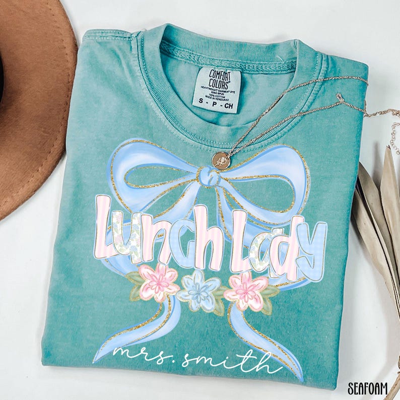 Puede incluir: Camiseta verde mar con un lazo azul claro y las palabras "Lunch Lady" en una fuente pastel. La camiseta tambi&eacute;n presenta detalles florales y el nombre "mrs. smith" en cursiva. Un collar plateado con un colgante est&aacute; sobre la camiseta.