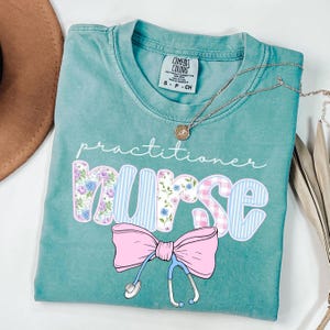 May include: A teal blue t-shirt with the text "practitioner nurse" in white. The word "nurse" is decorated with floral and plaid patterns. A pink bow with a stethoscope is below the text.