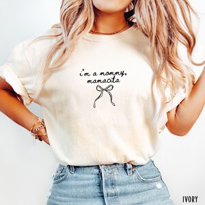 May include: Ivory-colored t-shirt with the text "i'm a mommy, mamacita" in a handwritten font, and a bow graphic. The shirt is made of a soft material and has rolled-up sleeves. The word "IVORY" is printed at the bottom right.