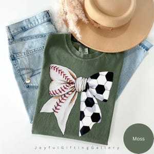 May include: A green t-shirt with a white bow with a baseball and soccer ball design. The text "Joyful Gifting Gallery" is at the bottom of the shirt. The color of the shirt is "Moss".