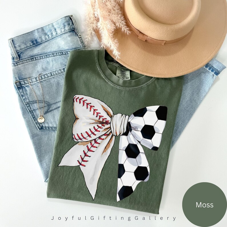 May include: A green t-shirt with a white bow with a baseball and soccer ball design. The text "Joyful Gifting Gallery" is at the bottom of the shirt. The color of the shirt is "Moss".