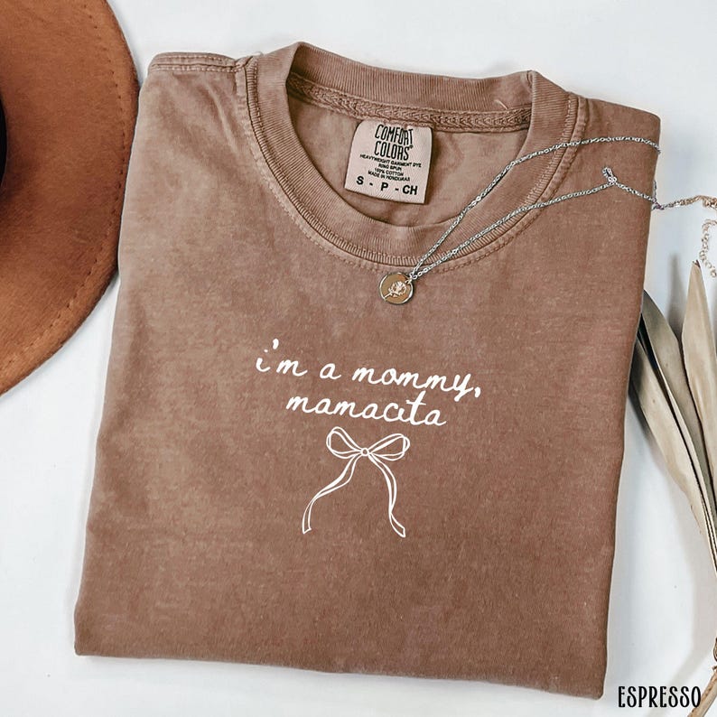 May include: Espresso-colored t-shirt with the text "I'm a mommy, mamacita" and a bow graphic. The shirt has a crew neck and a small tag on the upper back. A silver necklace with a pendant is draped across the shirt.