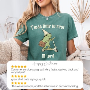 May include: Light green t-shirt with the text "T'was time to rest M'lord". The shirt features a cartoon frog riding a scooter and wearing a wizard hat. The shirt is light green.