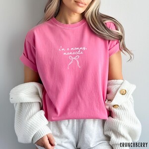 May include: A bright pink t-shirt with the text "I'm a mommy, mamacita" in white script, along with a bow graphic. The shirt has short sleeves and a crew neck.