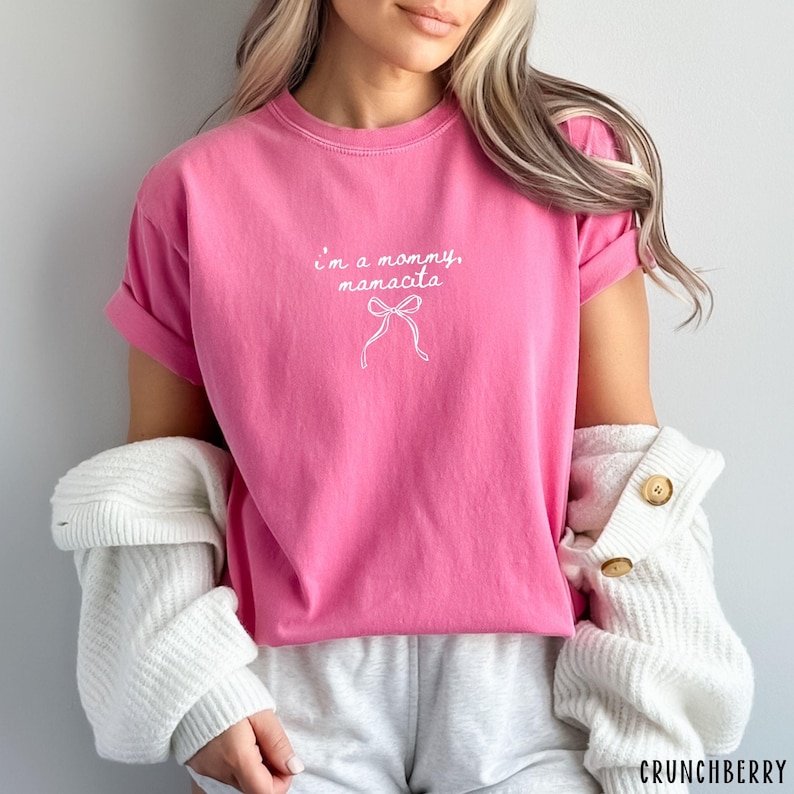 May include: A bright pink t-shirt with the text "I'm a mommy, mamacita" in white script, along with a bow graphic. The shirt has short sleeves and a crew neck.