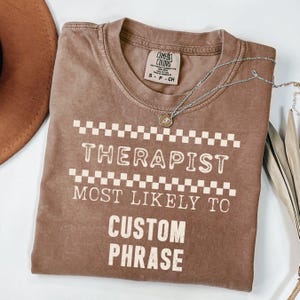 May include: A brown Comfort Colors t-shirt with the text "THERAPIST MOST LIKELY TO CUSTOM PHRASE" in white. The shirt has a checkerboard design above and below the word "THERAPIST". A silver necklace with a small pendant is draped across the shirt.