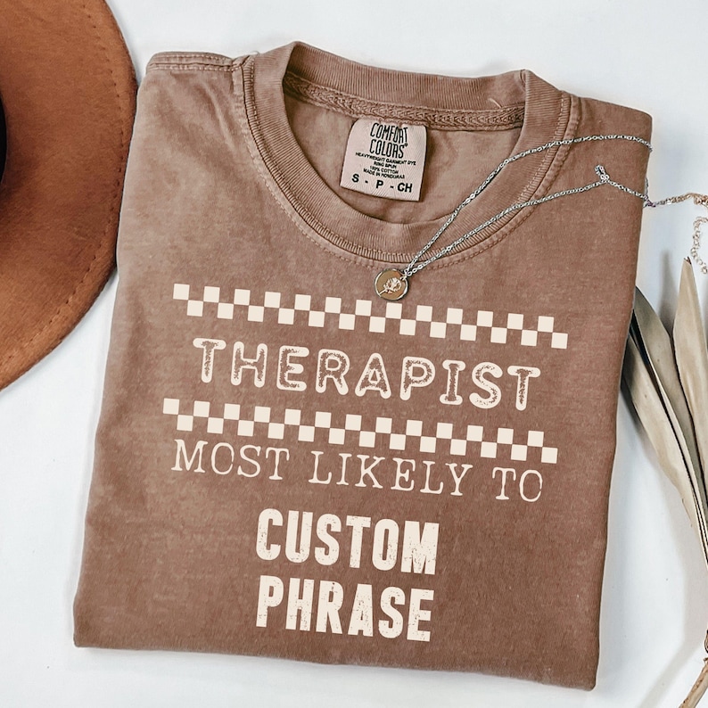 May include: A brown Comfort Colors t-shirt with the text "THERAPIST MOST LIKELY TO CUSTOM PHRASE" in white. The shirt has a checkerboard design above and below the word "THERAPIST". A silver necklace with a small pendant is draped across the shirt.