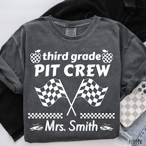 May include: A charcoal gray t-shirt featuring white screen-printed graphics. The design includes the text "third grade PIT CREW" with two checkered flags, two checkered apples, a checkered border, and the name "Mrs. Smith" with race car icons.