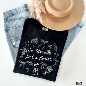 May include: Black Comfort Colors t-shirt with the text "I'm literally just a florist" surrounded by floral illustrations. The shirt is paired with light blue jeans, a tan hat, and a gold necklace.