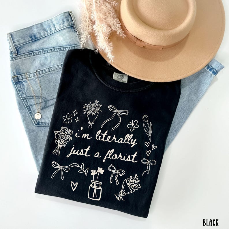 May include: Black Comfort Colors t-shirt with the text "I'm literally just a florist" surrounded by floral illustrations. The shirt is paired with light blue jeans, a tan hat, and a gold necklace.