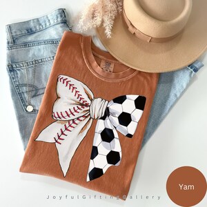 May include: A brown t-shirt with a white and black bow made from a baseball and a soccer ball. The bow is tied in the center of the shirt. The shirt is on a light blue background. The color of the shirt is called "Yam".