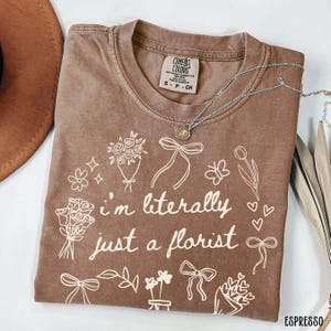 May include: Espresso-colored t-shirt with the text "I'm literally just a florist" and floral illustrations. The shirt has a crew neck and short sleeves. A silver necklace with a pendant is draped across the shirt.