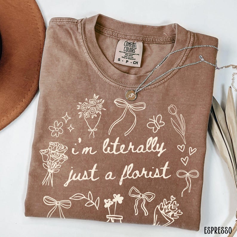 May include: Espresso-colored t-shirt with the text "I'm literally just a florist" and floral illustrations. The shirt has a crew neck and short sleeves. A silver necklace with a pendant is draped across the shirt.