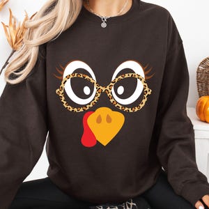 May include: A dark brown sweatshirt featuring a whimsical turkey face design. The turkey has large white eyes, brown eyelashes, and is wearing leopard print glasses. The beak is yellow and the wattle is red. A fun and festive design for Thanksgiving.