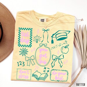 May include: A pale yellow t-shirt featuring a Comfort Colors label and various teal and light purple graphics including a graduation cap, an apple, a book, bows, and the text "test day," "you got this," "don't stress," and "do your best."