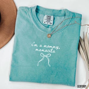 May include: Seafoam green t-shirt with the text "I'm a mommy, mamacita" and a bow graphic. A gold necklace with a pendant is draped across the shirt. The shirt is labeled "Comfort Colors" and is a size small.