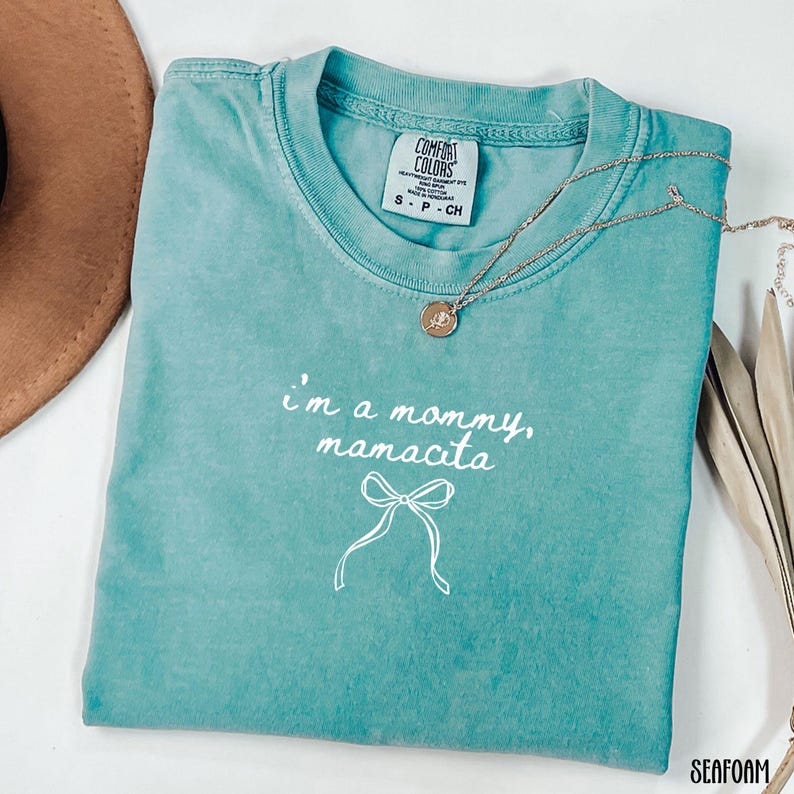 May include: Seafoam green t-shirt with the text "I'm a mommy, mamacita" and a bow graphic. A gold necklace with a pendant is draped across the shirt. The shirt is labeled "Comfort Colors" and is a size small.
