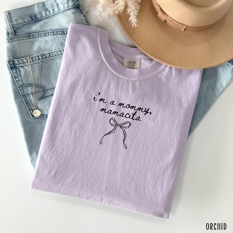 May include: A light purple t-shirt with the text "I'm a mommy, mamacita" in black script, along with a bow graphic. The shirt is folded on top of blue jeans, with a gold necklace and a tan hat visible.