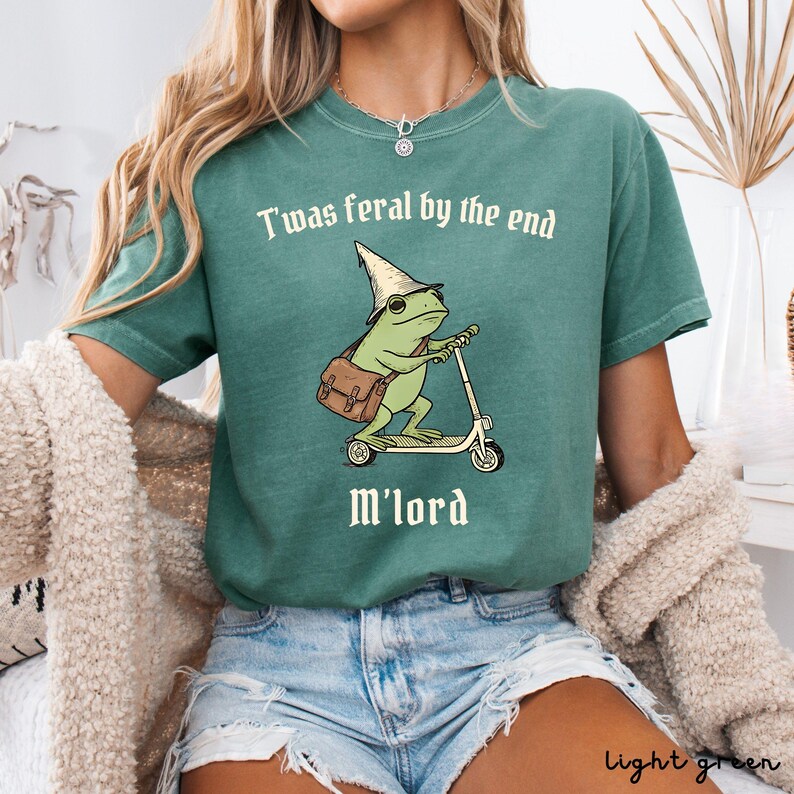 May include: Light green t-shirt with the text "T'was feral by the end M'lord." The shirt features a cartoon frog wearing a wizard hat and riding a scooter. The frog has a brown satchel bag.