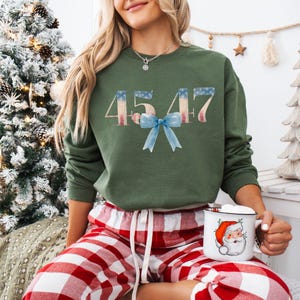 May include: A green sweatshirt with the numbers 4547 printed in red, white, and blue, resembling the American flag. A blue bow is centered below the numbers. The sweatshirt is worn by a person wearing red and white plaid pajama pants.