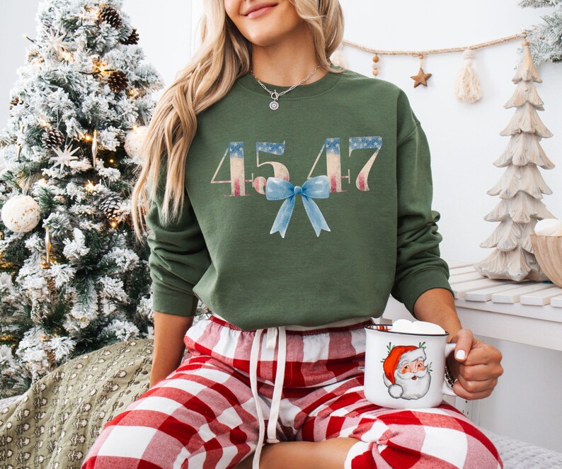May include: A green sweatshirt with the numbers 4547 printed in red, white, and blue, resembling the American flag. A blue bow is centered below the numbers. The sweatshirt is worn by a person wearing red and white plaid pajama pants.