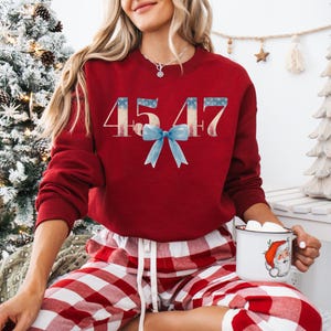 May include: Red sweatshirt with the number 4547 printed in white with a blue bow. The sweatshirt is worn by a person wearing red and white plaid pajama pants.